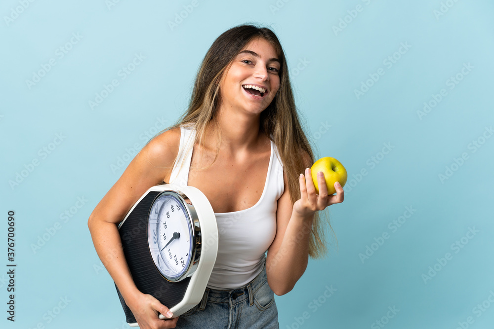 Young caucasian woman isolated on blue background with weighing machine and with an apple Young caucasian woman isolated on blue background with weighing machine and with an apple