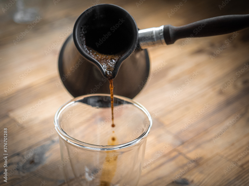 Freshly brewed turkish coffee pouring from cezve into transparent glass on wooden background. Filling hot coffee to transparent vacuum glass with double walls. Freshly brewed turkish coffee pouring from cezve into transparent glass on wooden background. Filling hot coffee to transparent vacuum glass with double walls.
