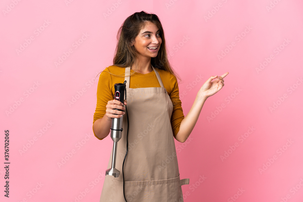 Young chef woman using hand blender isolated on pink background pointing finger to the side Young chef woman using hand blender isolated on pink background pointing finger to the side