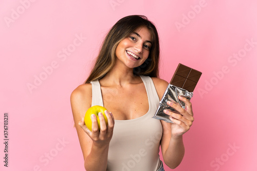 Young caucasian woman isolated on pink background taking a chocolate tablet in one hand and an apple in the other
