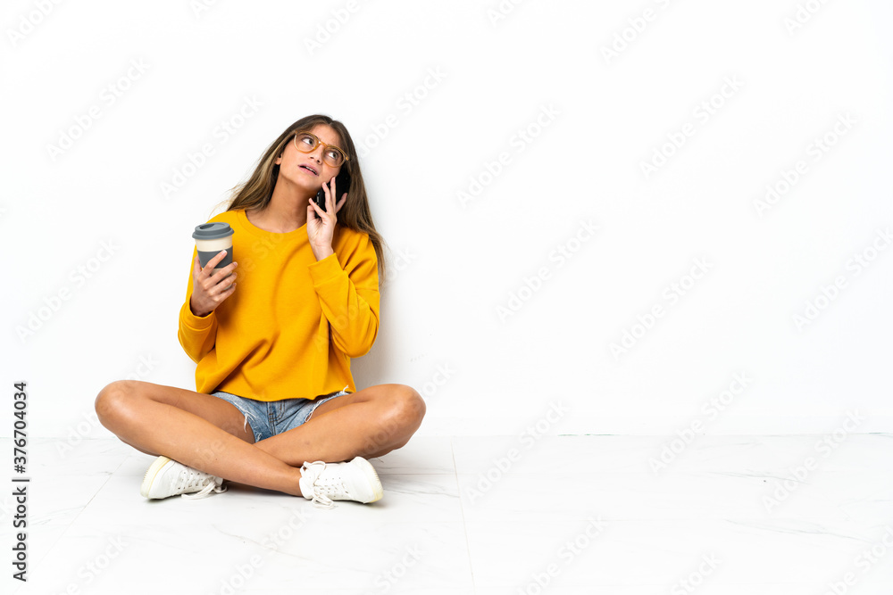 Young woman sitting on the floor isolated on white background holding coffee to take away and a mobile