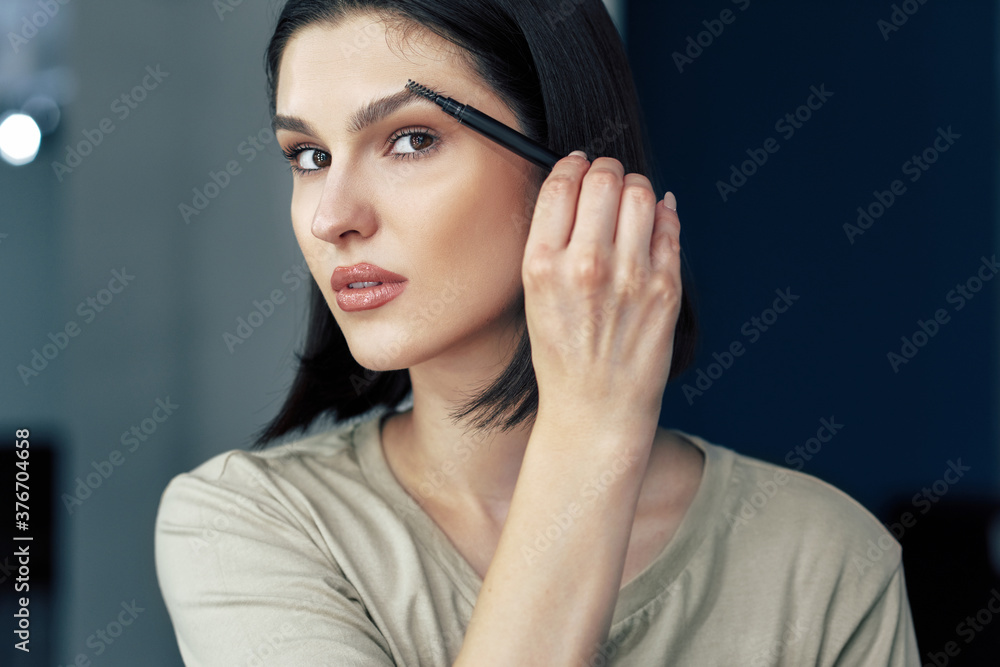 Portrait of beautiful makeup artist girl applying gel for eyebrow in front of mirror. Young brunette woman making herself make-up while recording her video for her blog on cosmetics. Portrait of beautiful makeup artist girl applying gel for eyebrow in front of mirror. Young brunette woman making herself make-up while recording her video for her blog on cosmetics.