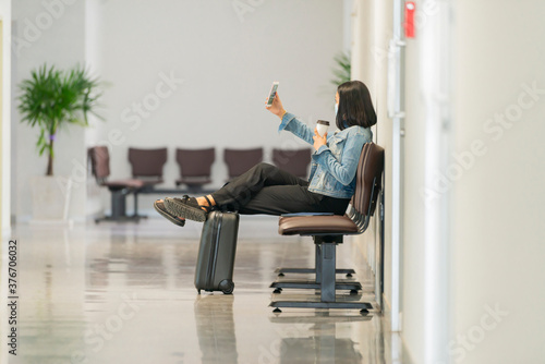Young asian businesswoman is photographing herself on cell telephone while is relaxing with take away coffee in the departure hall at airport. Copy space