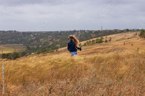 Adorable little girl in denim jacket, blue plaid dress running in yellow wild grass field. Happy stylish long blonde hair child on countryside landscape. Cute kid walking outdoor rural road trip.