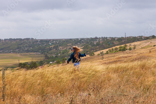 Adorable little girl in denim jacket, blue plaid dress running in yellow wild grass field. Happy stylish long blonde hair child on countryside landscape. Cute kid walking outdoor rural road trip. © KawaiiS