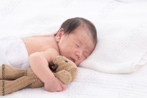 Asian adorable 0-10 day newborn baby boy sleeping comfortable in bed hold bear toy, cute infant lying down on white bed at home with copy space, 0-10 day happy childhood sleep in bed concept