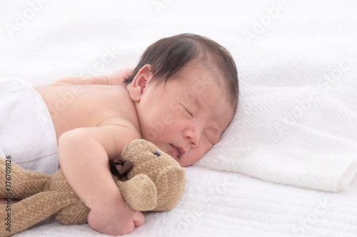 Asian adorable 0-10 day newborn baby boy sleeping comfortable in bed hold bear toy, cute infant lying down on white bed at home with copy space, 0-10 day happy childhood sleep in bed concept