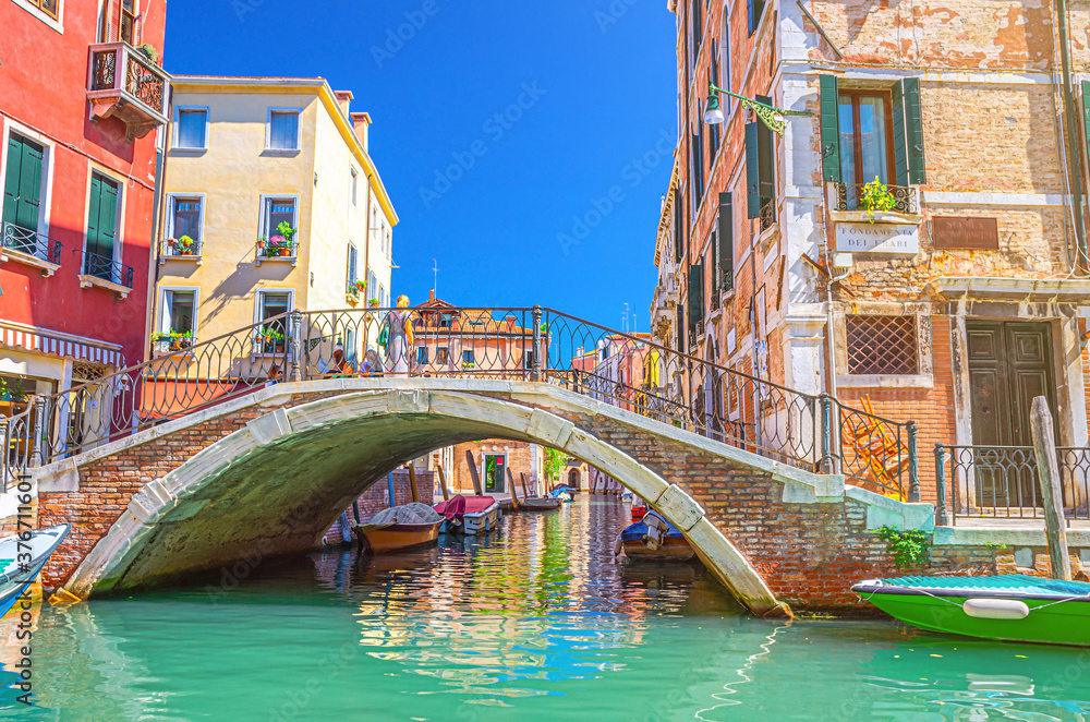 Bridge across narrow water canal in Venice with moored boats between old colorful buildings with balconies and brick walls, blue sky, Veneto Region, Northern Italy. Typical Venetian cityscape