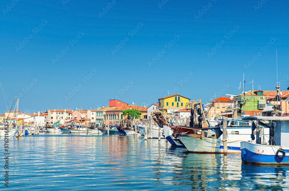 Fishing ships and boats moored in port lagoon near seaside of Sottomarina town with row of colorful buildings in summer day, blue sky background, Veneto Region, Northern Italy