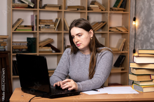 young woman looking at a laptop at her desk with books and documents. bookcase in the background in the office or at home. student on online education. remote work in quarantine.