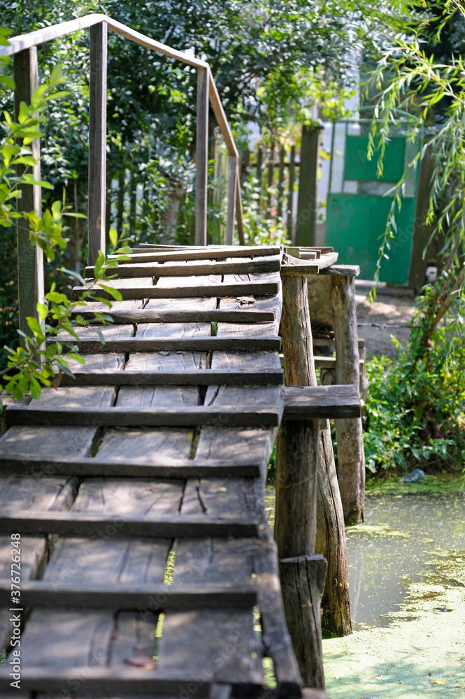 An old wooden footbridge built on the canal, household on a background An old wooden footbridge built on the canal, household on a background