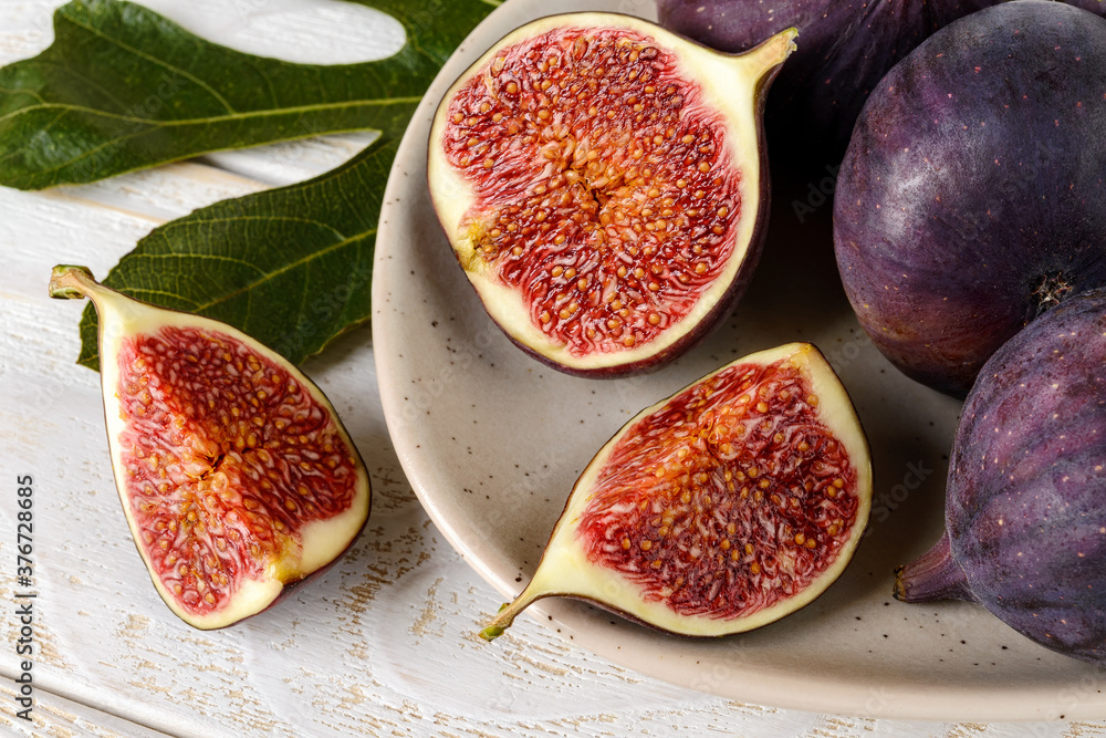 Close-up of fresh ripe fig fruits on a beige ceramic plate and a green leaf over a white wood table. Sweet fruits and berries. Vegetarian, raw food diet and healthy eating. Close-up of fresh ripe fig fruits on a beige ceramic plate and a green leaf over a white wood table. Sweet fruits and berries. Vegetarian, raw food diet and healthy eating.