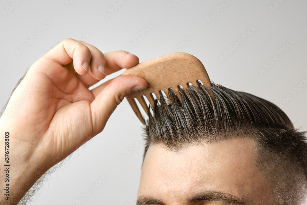 Close up man brushing his hair with wooden comb on the grey background. Treatment against hair lost and dandruff concept. Cosmetic products for men. Barbershop advertising.  - obrazy, fototapety, plakaty 
