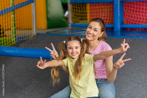 Cheerful mother and her daughter makign peace gesture at indoor kids playground, empty space photo