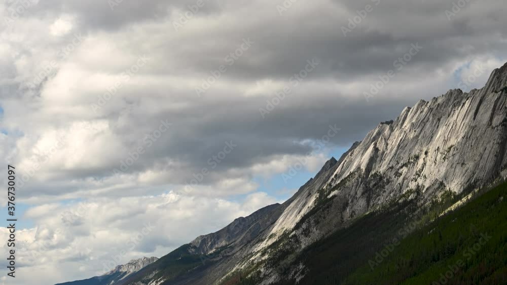 Dramatic clouds move through the sky above a large grey mountain cliff in this time lapse. Green trees occur at the base of the mountain.
Dramatic clouds move through the sky above a large grey mountain cliff in this time lapse. Green trees occur at the base of the mountain.