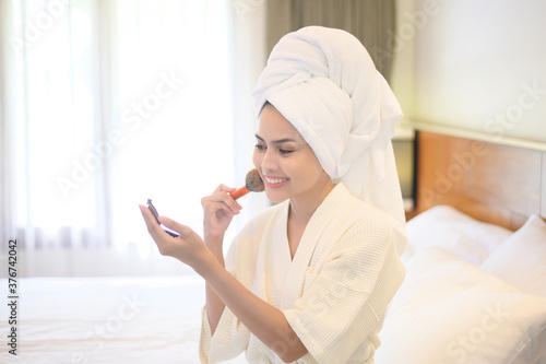 Attractive happy female in white bathrobe is applying natural Make-Up with cosmetic powder brush, Beauty Concept.