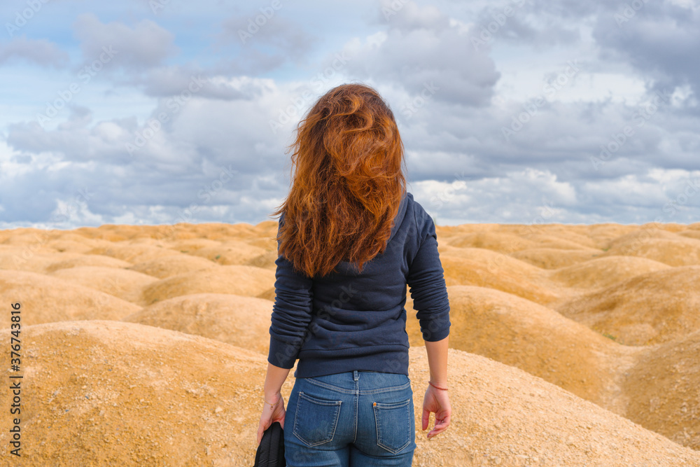 A young woman with red hair stands with her back to the camera in a desert with sand dunes of bizarre shape