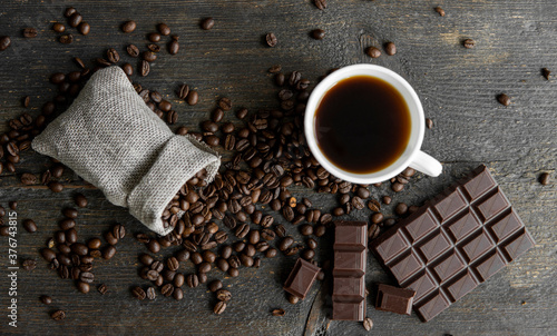 Coffee beans scattered from a linen bag on a wooden table. Cup of coffee and bar of dark chocolate. Mug of black coffee. Morning coffee with a chocolate.