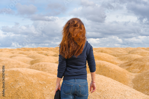 A young woman with red hair stands with her back to the camera in a desert with sand dunes of bizarre shape © KseniaJoyg