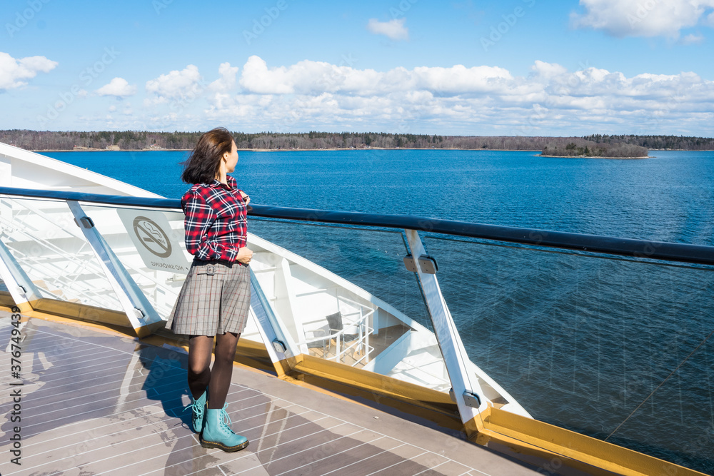 Fototapeta premium Beautiful slender girl traveler on the upper deck of cruise ship. Asian young woman dressed in red plaid shirt and short skirt in luxury cruise. Elegant happy woman look at ocean relax on luxury liner