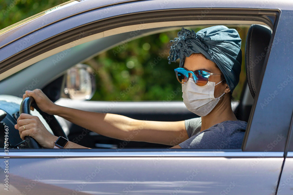 A woman driving a car and wearing a mask, scarf in her hair and sunglasses to protect herself from contamination by corona virus. Pandemic. Social isolation. Essential services.
