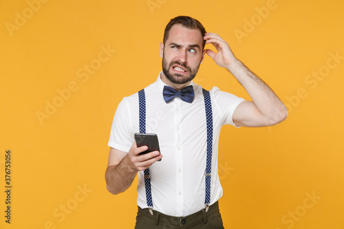 Preoccupied young bearded man 20s wearing white shirt bow-tie suspender posing using mobile cell phone typing sms message put hand on head isolated on bright yellow color background studio portrait.