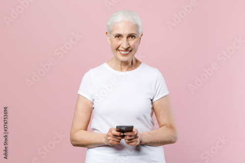 Smiling elderly gray-haired female woman wearing white casual t-shirt posing hold mobile cell phone typing sms message looking camera isolated on pastel pink color wall background studio portrait. © ViDi Studio
