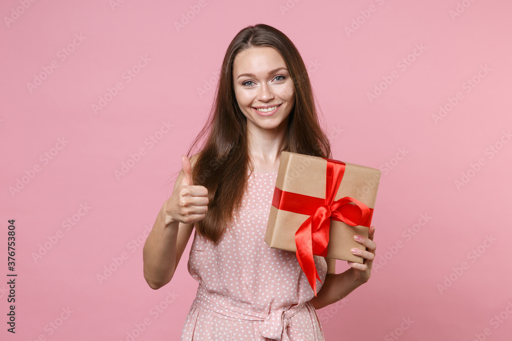 Smiling young brunette woman wearing pink summer dotted dress posing celebrating hold red present box with gift ribbon bow showing thumb up isolated on pastel pink color background studio portrait. Smiling young brunette woman wearing pink summer dotted dress posing celebrating hold red present box with gift ribbon bow showing thumb up isolated on pastel pink color background studio portrait.