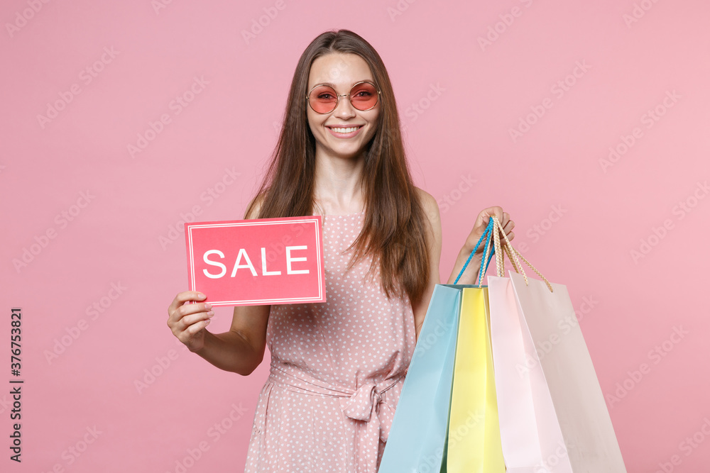 Smiling young brunette woman in pink summer dotted dress glasses hold package bag with purchases after shopping sign with SALE title looking camera isolated on pastel pink background studio portrait. Smiling young brunette woman in pink summer dotted dress glasses hold package bag with purchases after shopping sign with SALE title looking camera isolated on pastel pink background studio portrait.