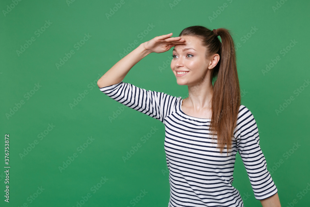Smiling beautiful young brunette woman 20s wearing striped casual clothes posing standing holding hand at forehead looking far away distance isolated on green color wall background, studio portrait. Smiling beautiful young brunette woman 20s wearing striped casual clothes posing standing holding hand at forehead looking far away distance isolated on green color wall background, studio portrait.