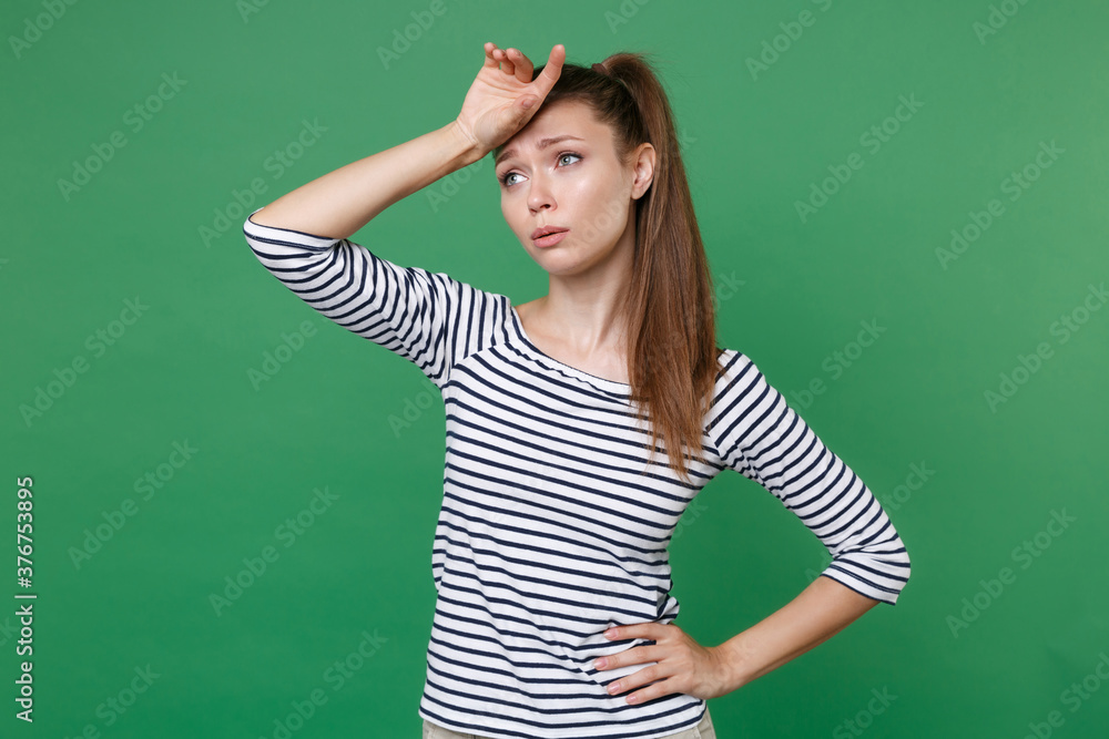 Exhausted tired displeased dissatisfied puzzled young brunette woman 20s wearing striped casual clothes posing put hand on head looking aside up isolated on green color background studio portrait.
