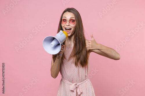 Surprised excited young brunette woman 20s wearing pink summer dotted dress eyeglasses screaming in megaphone showing thumb up looking camera isolated on pastel pink color background studio portrait.