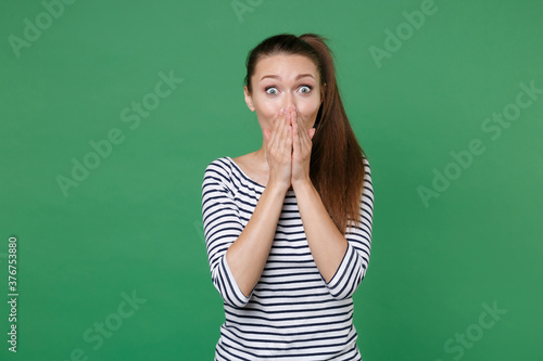 Shocked amazed surprised excited young brunette woman 20s wearing striped casual clothes posing standing covering mouth with hands looking camera isolated on green color background, studio portrait.