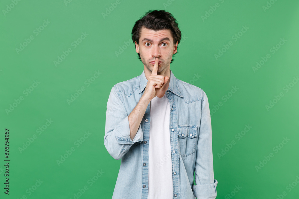 Secret young brunet man 20s wearing casual clothes white t-shirt denim shirt posing saying hush be quiet with finger on lips shhh gesture looking camera isolated on green background studio portrait.