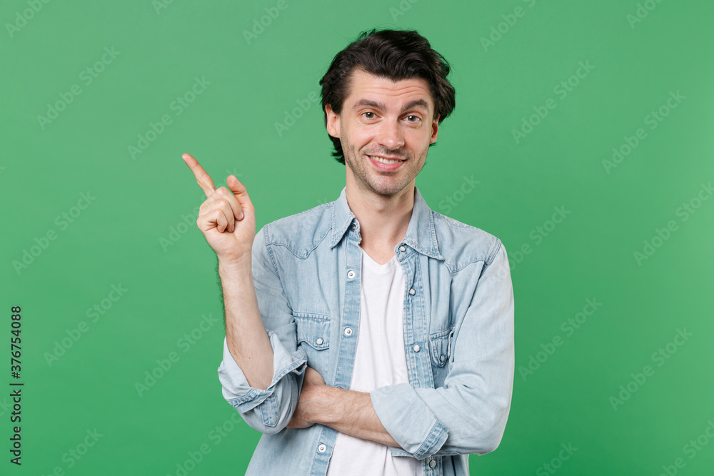 Smiling young brunet man 20s wearing casual clothes white t-shirt denim shirt posing standing pointing index finger aside up on mock up copy space isolated on green color background studio portrait.