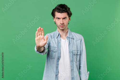 Tableau sur toile Serious young brunet man 20s wearing casual clothes white t-shirt denim shirt posing standing showing stop gesture with palm looking camera isolated on green color wall background studio portrait
