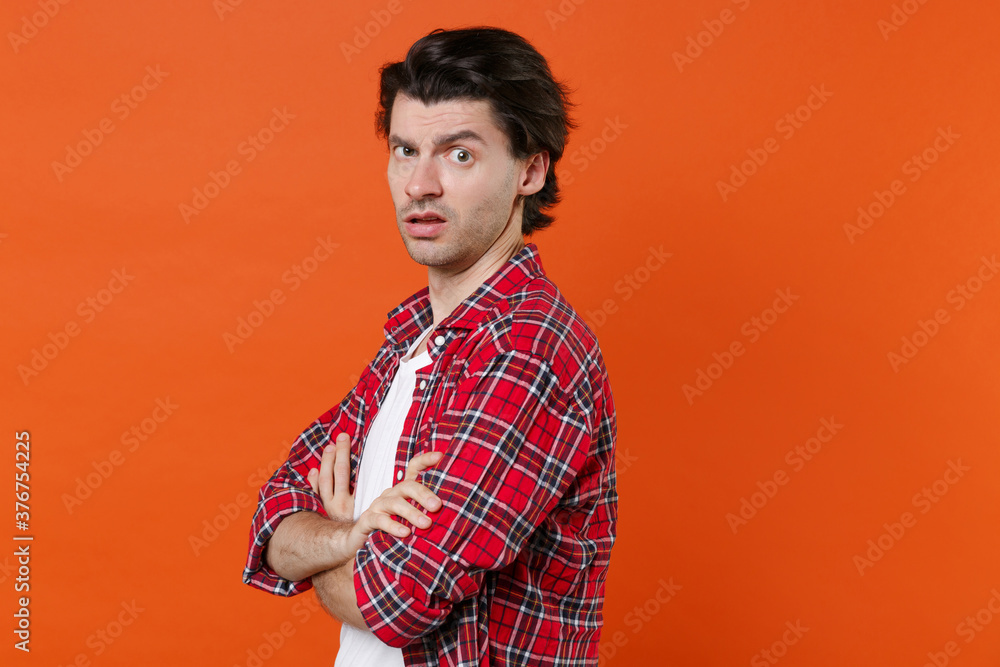 Side view of perplexed puzzled young brunet man 20s wearing white t-shirt red checkered shirt posing holding hands crossed looking camera isolated on orange color wall background studio portrait. Side view of perplexed puzzled young brunet man 20s wearing white t-shirt red checkered shirt posing holding hands crossed looking camera isolated on orange color wall background studio portrait.