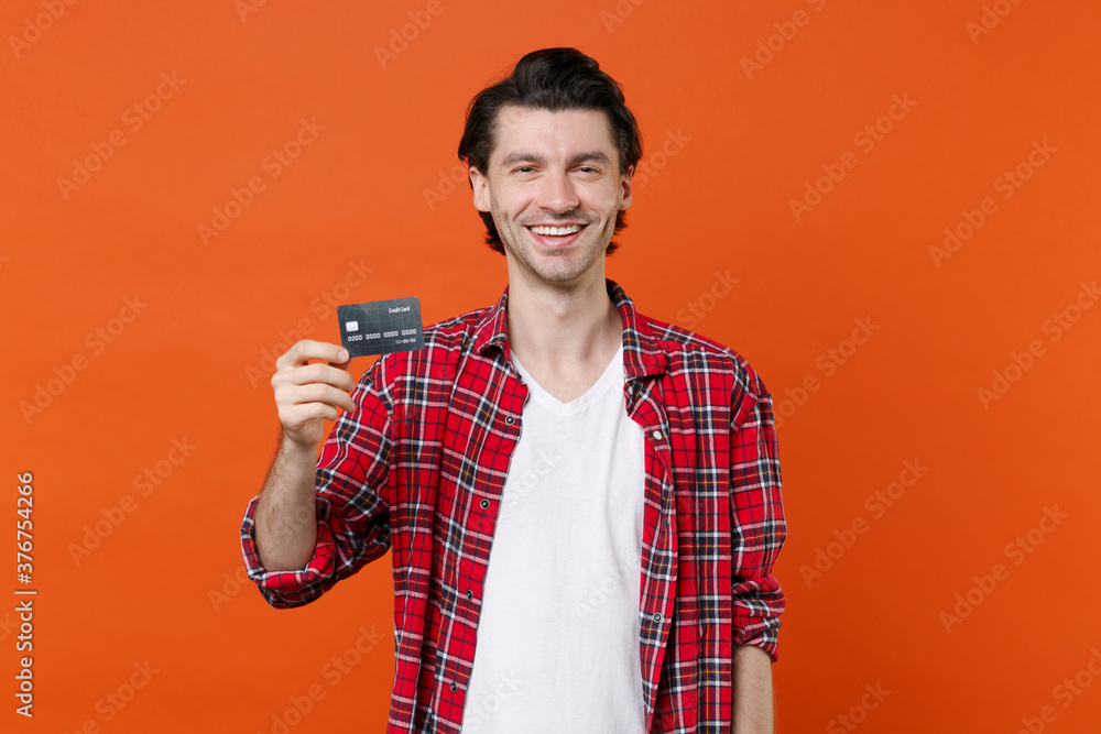 Smiling cheerful young brunet man 20s in casual clothes white t-shirt red checkered shirt posing standing hold in hands credit bank card looking camera isolated on orange background studio portrait. Smiling cheerful young brunet man 20s in casual clothes white t-shirt red checkered shirt posing standing hold in hands credit bank card looking camera isolated on orange background studio portrait.