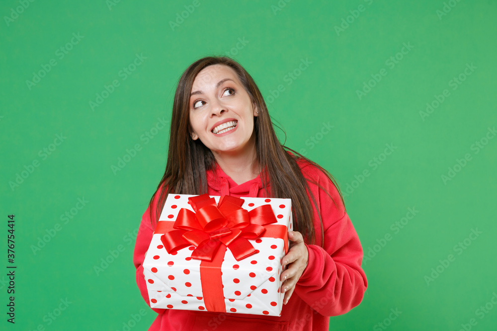 Smiling pretty pensive young brunette woman in red casual streetwear hoodie posing celebrating hold red present box with gift ribbon bow looking up isolated on green color background studio portrait. Smiling pretty pensive young brunette woman in red casual streetwear hoodie posing celebrating hold red present box with gift ribbon bow looking up isolated on green color background studio portrait.