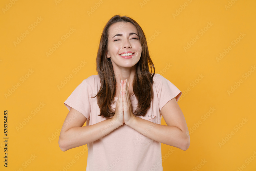 Pleading smiling young brunette woman 20s wearing pastel pink casual t-shirt posing holding hands folded in prayer keeping eyes closed isolated on bright yellow color wall background studio portrait. Pleading smiling young brunette woman 20s wearing pastel pink casual t-shirt posing holding hands folded in prayer keeping eyes closed isolated on bright yellow color wall background studio portrait.
