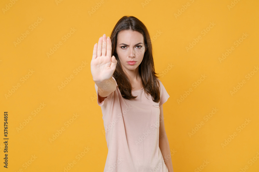 Displeased dissatisfied young brunette woman wearing pastel pink casual t-shirt standing posing showing stop gesture with palm looking camera isolated on yellow color wall background studio portrait. Displeased dissatisfied young brunette woman wearing pastel pink casual t-shirt standing posing showing stop gesture with palm looking camera isolated on yellow color wall background studio portrait.