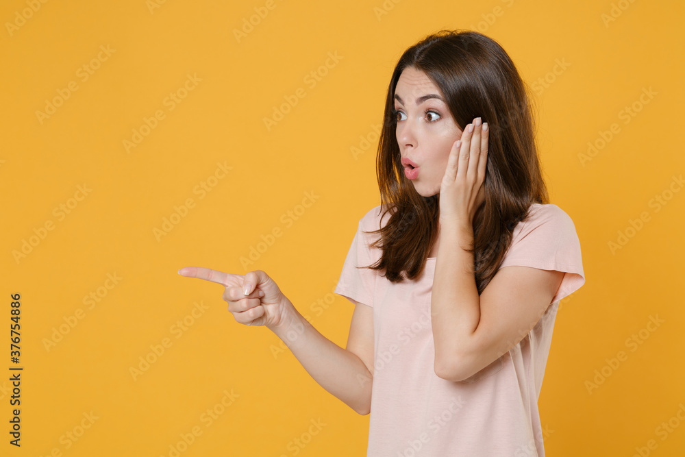 Shocked young brunette woman 20s in pastel pink casual t-shirt standing posing pointing index finger aside on mock up copy space put hand on cheek isolated on yellow color background studio portrait. Shocked young brunette woman 20s in pastel pink casual t-shirt standing posing pointing index finger aside on mock up copy space put hand on cheek isolated on yellow color background studio portrait.