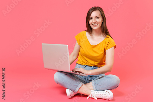 Full length portrait of beautiful smiling young brunette woman 20s wearing yellow t-shirt sitting on floor working on laptop pc computer looking camera isolated on pink color wall background studio.
