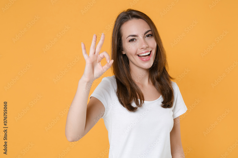 Cheerful funny beautiful attractive young woman 20s wearing white blank empty design casual t-shirt posing standing showing OK gesture isolated on yellow color wall background studio portrait. Cheerful funny beautiful attractive young woman 20s wearing white blank empty design casual t-shirt posing standing showing OK gesture isolated on yellow color wall background studio portrait.