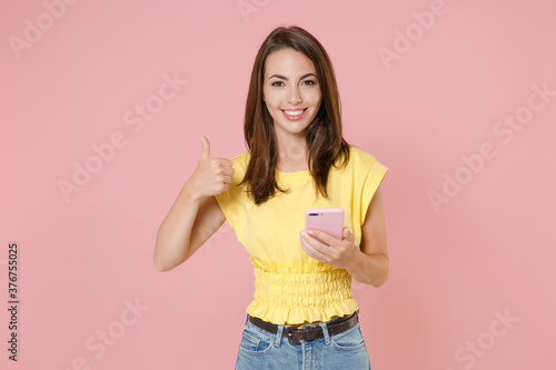 Smiling young brunette woman 20s wearing yellow casual t-shirt posing standing using mobile cell phone typing sms message showing thumb up isolated on pastel pink color background studio portrait.