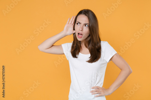Shocked curious amazed young woman 20s in white blank empty design casual t-shirt posing try to hear you overhear listening intently looking aside isolated on yellow color background studio portrait.