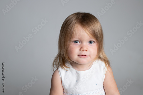 Portrait of a cute little girl 1.5-2 years old. Blonde, blue eyes, shoulder hair. He carefully looks at the viewer, smiles. The face of a healthy child. Studio lighting, horizontal. Positive baby.