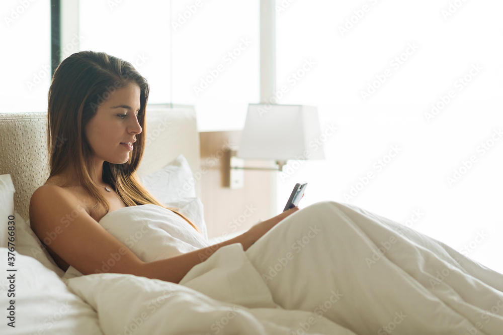 Young woman sitting up in hotel bed reading smartphone texts Young woman sitting up in hotel bed reading smartphone texts