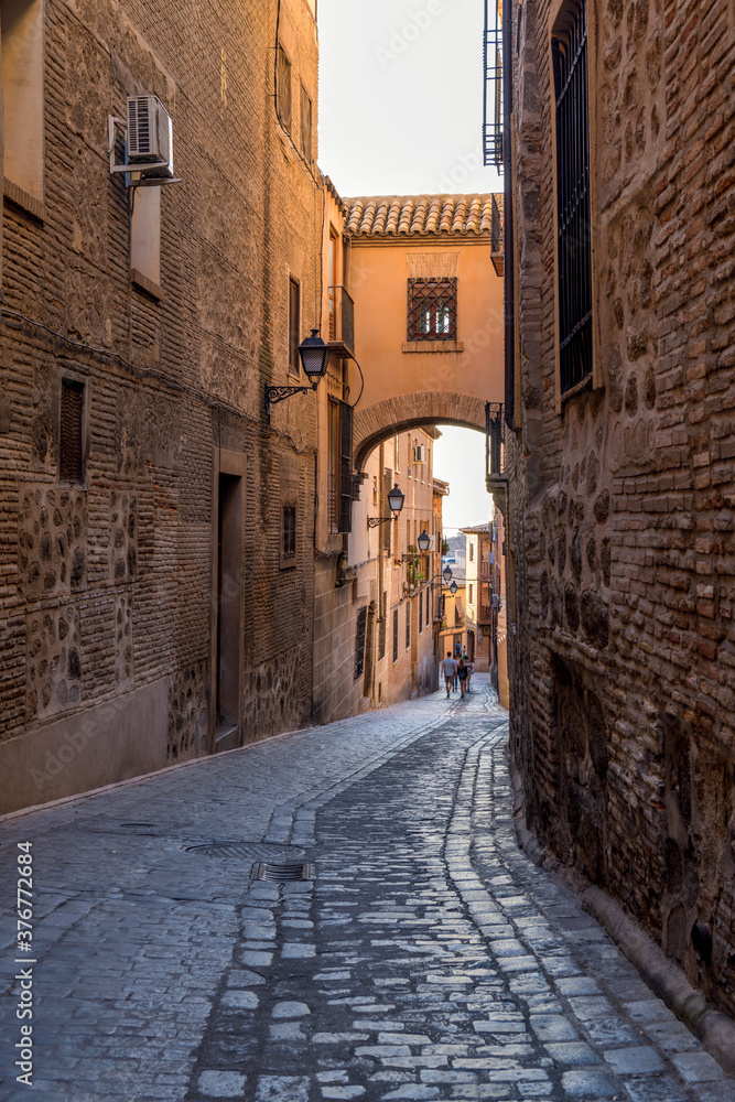 Cobblestone Street - A sunny evening view of a brick bridge above an old narrow cobblestone street in Jewish Quarter of the historic city Toledo, Spain. - obrazy, fototapety, plakaty 