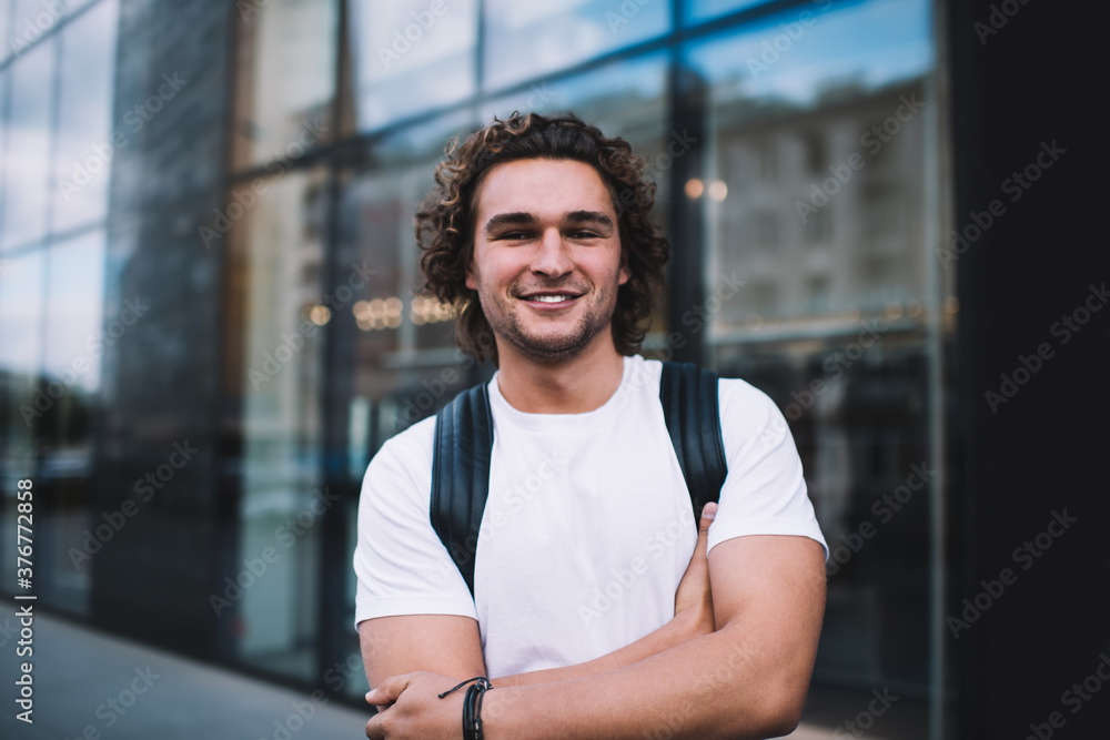 Half length portrait of handsome smiling hipster guy posing on urban background with crossed hands, happy male traveler in white shirt with copy space for brand name or label outdoors look at camera